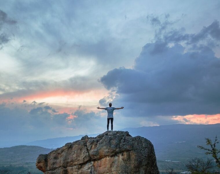 Hombre de pie sobre un monte con las manos elevadas al un cielo parcialmente nublado al anochecer