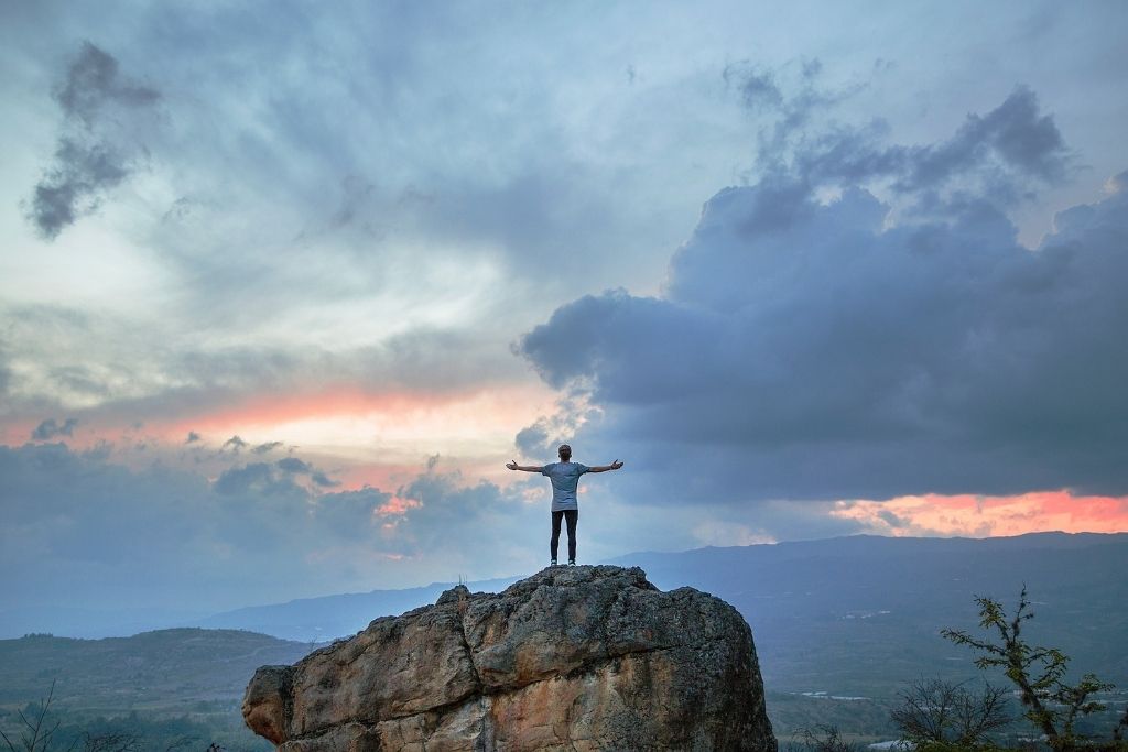 Hombre de pie sobre un monte con las manos elevadas al un cielo parcialmente nublado al anochecer